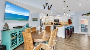 A modern kitchen with white cabinets, a wooden dining table, wicker chairs, and a large TV screen displaying a beach scene.