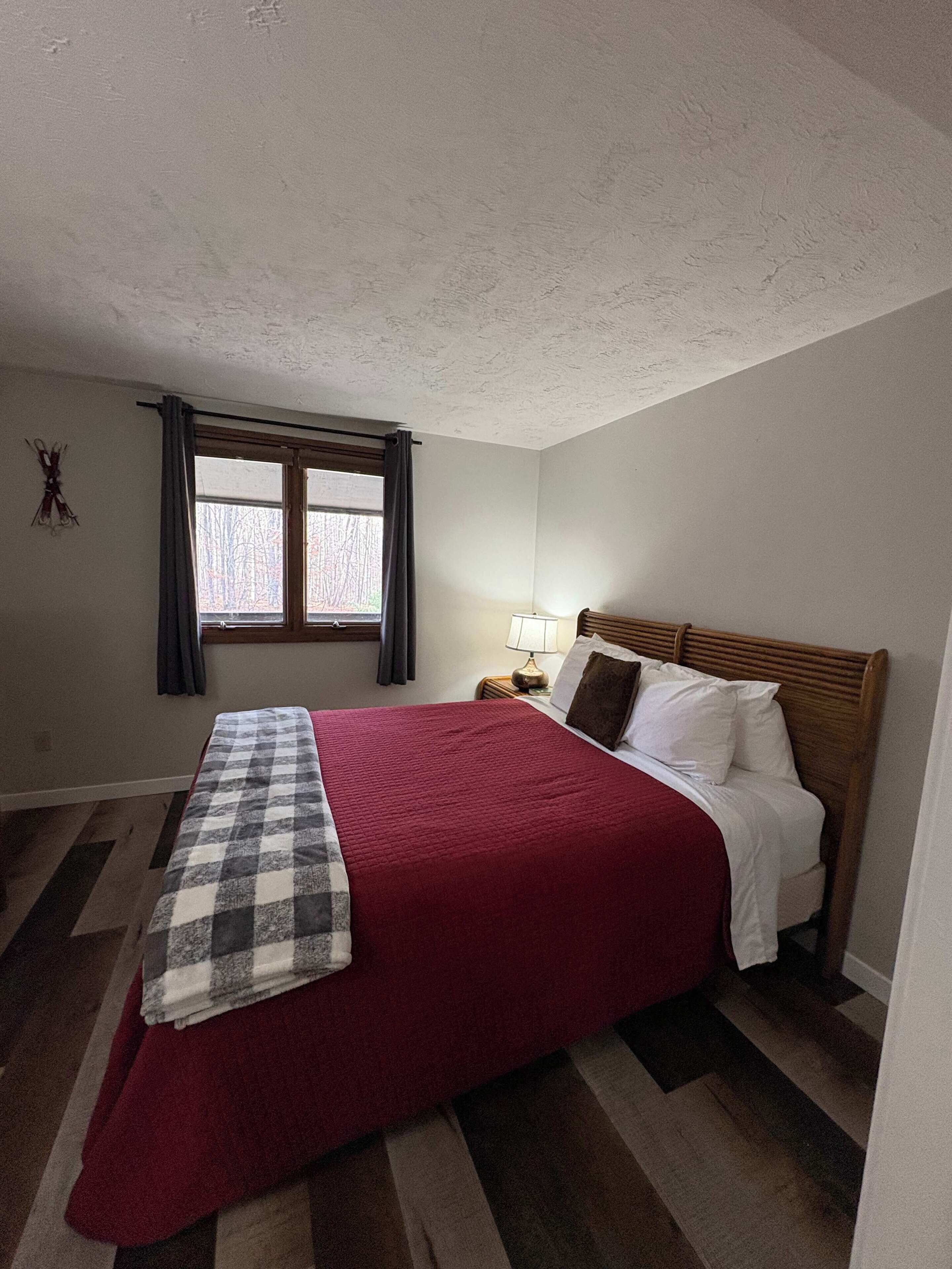 Ein Schlafzimmer mit Bett, einem Fenster mit Vorhängen, einer Nachttischlampe und einer karierten Decke.