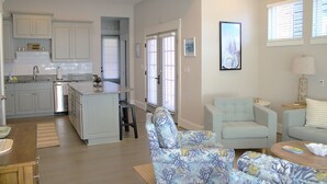 A spacious living area with a light blue armchair, a sofa, and a coffee table. There is a kitchen with white cabinets and a granite countertop. - 404A The Hilltop House | 4BR/3.5BA | Balcony (Virginia Beach)