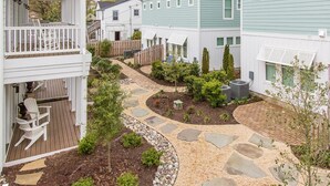 A residential area with a paved walkway, small trees, and a wooden deck with chairs. - 404A The Hilltop House | 4BR/3.5BA | Balcony (Virginia Beach)
