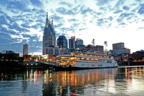 A riverfront cityscape with a prominent building featuring a pointed spire, a large white boat docked at the pier, and a reflection of the city lights on the water. - ❤️of DWTN on the RIVER/Walk 2 BROADWAY & ALL/❤️2KING BDS/2BA/Free PRKG/POOL!🎸🎻 (Nashville)