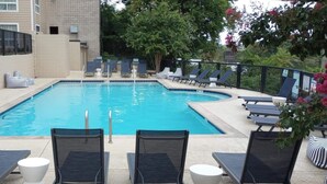 Une piscine rectangulaire, agrémentée de chaises longues et d’un espace dédié au bronzage, le tout entouré d’une clôture et d’arbres.