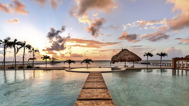 Une piscine avec un chemin de pierre menant à une structure au toit de chaume, entourée de palmiers et offrant une vue sur le coucher du soleil.