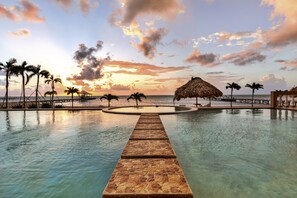 A swimming pool with a stone pathway leading to a thatched-roof structure, surrounded by palm trees and a sunset view.