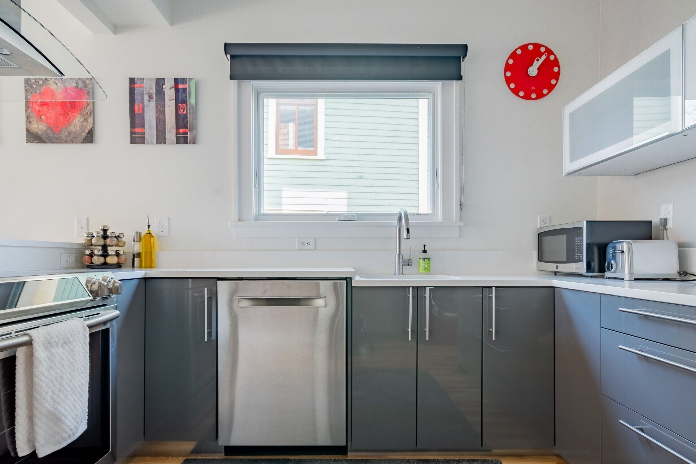 Une cuisine moderne équipée d’appareils électroménagers en acier inoxydable, d’un lave-vaisselle et d’un four à micro-ondes. On y trouve une fenêtre avec des stores, une horloge rouge et une petite fenêtre donnant sur l’extérieur.