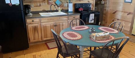 Een houten keuken met een zwarte koelkast, houten keukenkastjes, een spoelbak en een fornuis. Er staat een eettafel met vier stoelen en rode en witte placemats.