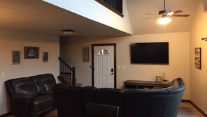A living room with a black leather sofa, a wooden coffee table, a flat-screen TV mounted on the wall, and a ceiling fan.