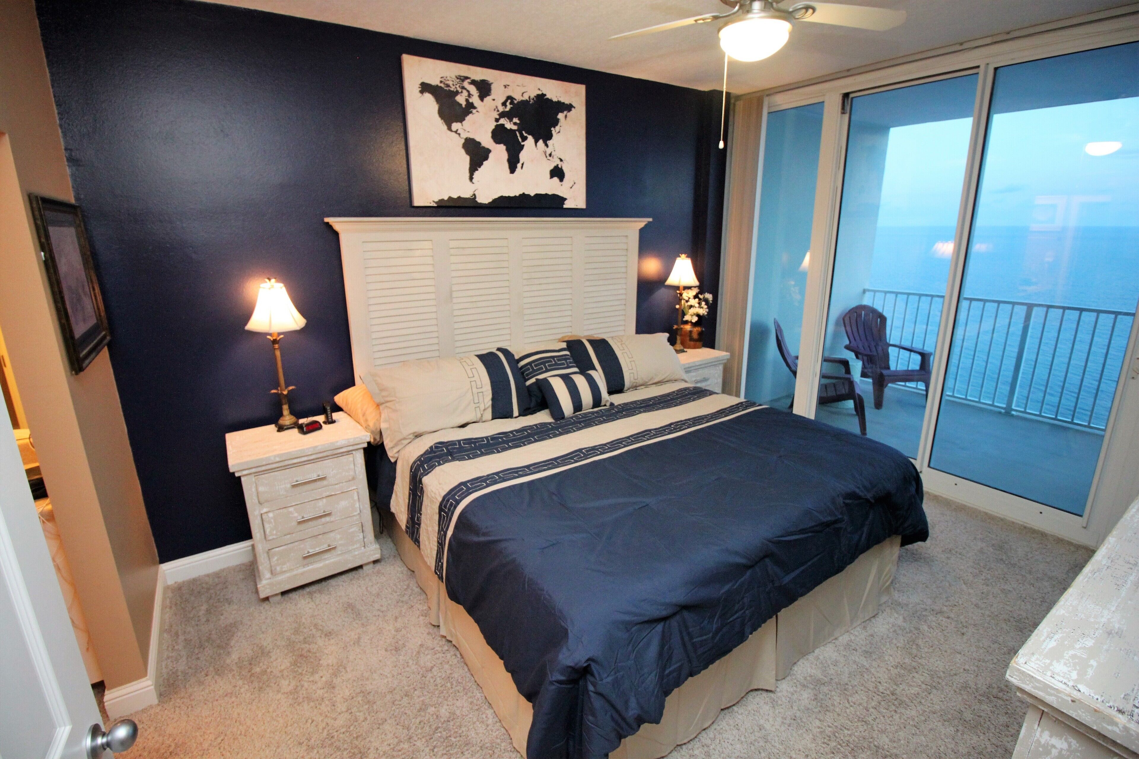 A bedroom with a large bed, two bedside tables with lamps, a world map on the wall, and a view of the ocean through a sliding glass door.