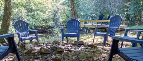 A campsite with several Adirondack chairs and picnic tables around a small pond, surrounded by trees.