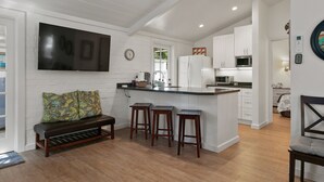 A modern kitchen with white cabinets, a central island with stools, a flat-screen TV mounted above the island, and a wooden bench with cushions in the adjacent living area. - Private Cottage! Easy Walk Through The Park To Encinitas And Moonlight Beach. (Encinitas)