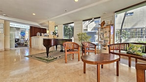 A spacious lobby with a grand piano, wooden chairs, and a coffee table. A reception desk is visible with a sign for real estate sales.