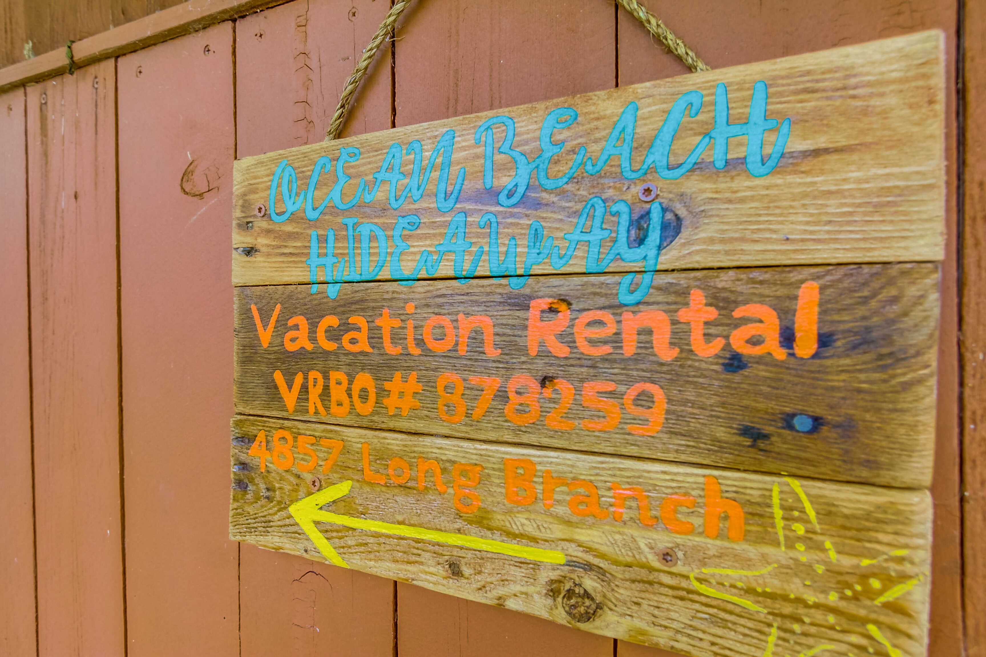Un letrero de madera con la frase 'Ocean Beach Hideaway' y 'Alquiler Vacacional', además de un número de teléfono y una flecha apuntando a la izquierda.