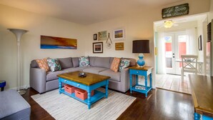 A living room with a grey sofa, a wooden coffee table, and a blue side table. There are framed pictures on the wall and a sign that says "Life is good at the beach."