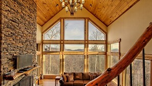 Interior acogedor de una cabaña con chimenea de piedra, un ventanal con vistas a la montaña y una escalera de madera.