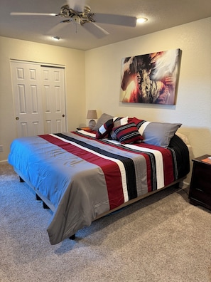A bedroom with a bed, a ceiling fan, a door, a wall-mounted lamp, and a painting.