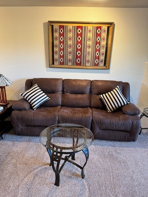A brown leather sofa with striped pillows, a glass coffee table, and a wall with a patterned tapestry.