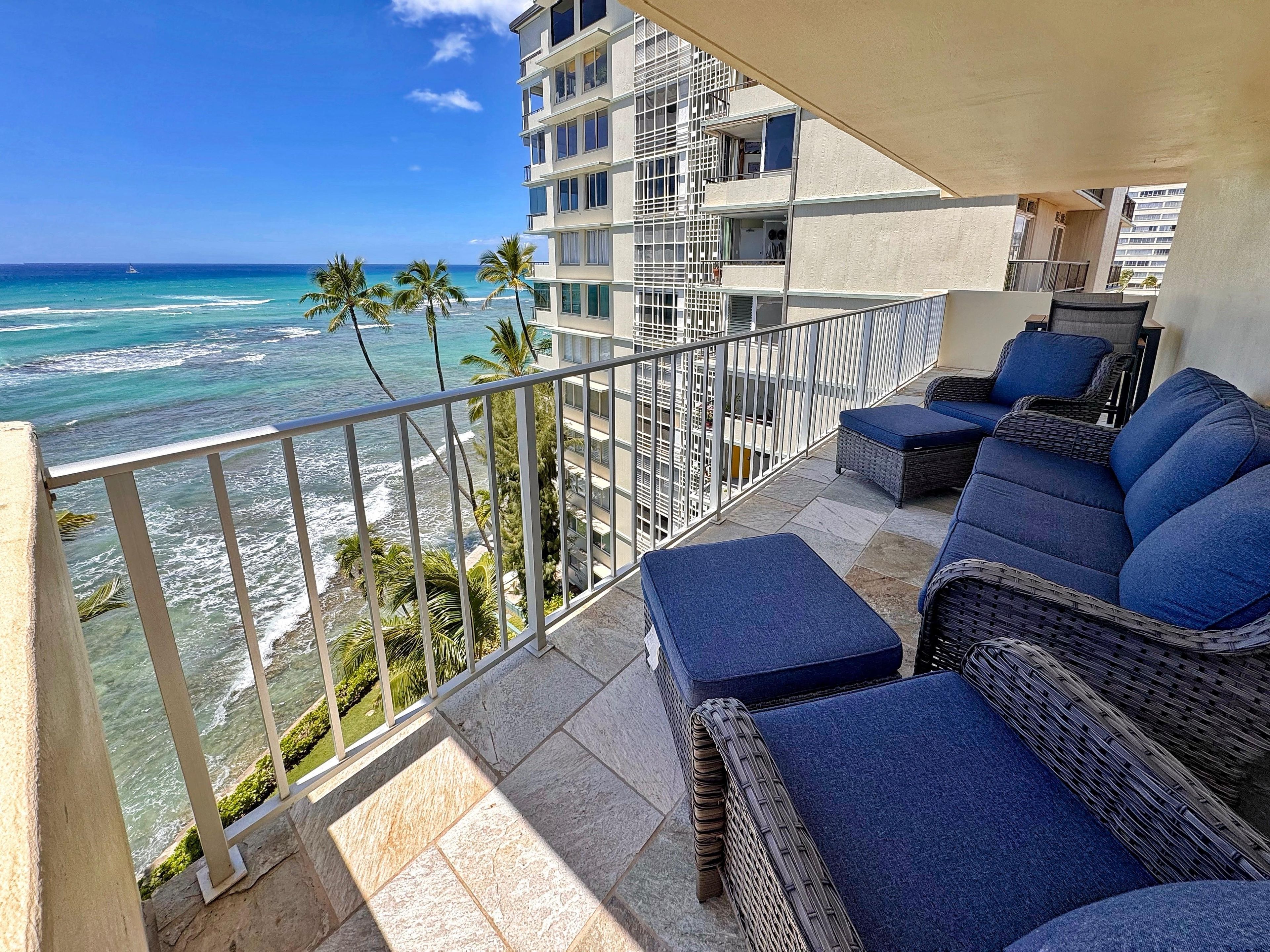 Balcony with slate tile flooring, dark wicker seating with blue cushions, and oceanfront views of palm trees and waves.