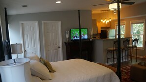 A bedroom with a bed, a television, a ceiling fan, and a view of a kitchen and dining area.