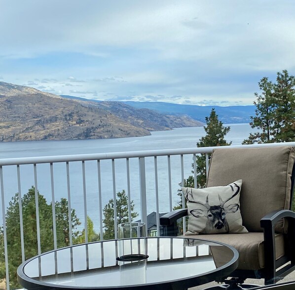 Un balcón con una mesa de vidrio y una silla, con vista a un lago y montañas.