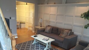 A living room with a gray sofa, a white coffee table, and a patterned rug. There is a staircase with wooden railings on the left. - Best Location In LBI! Walk To Everything (Beach Haven)