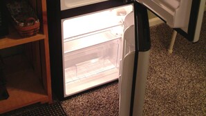 Un refrigerador compacto con un compartimento congelador superior, ubicado en un rincón de una habitación con ventana y estante de madera.