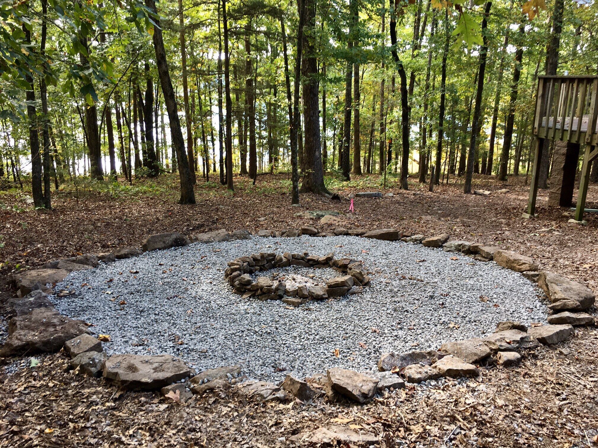 A circular gravel area surrounded by rocks, with a forested background and a wooden structure to the right.