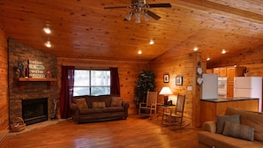 A cozy cabin interior with a fireplace, wooden floors, a sofa, a dining area, and a kitchen.