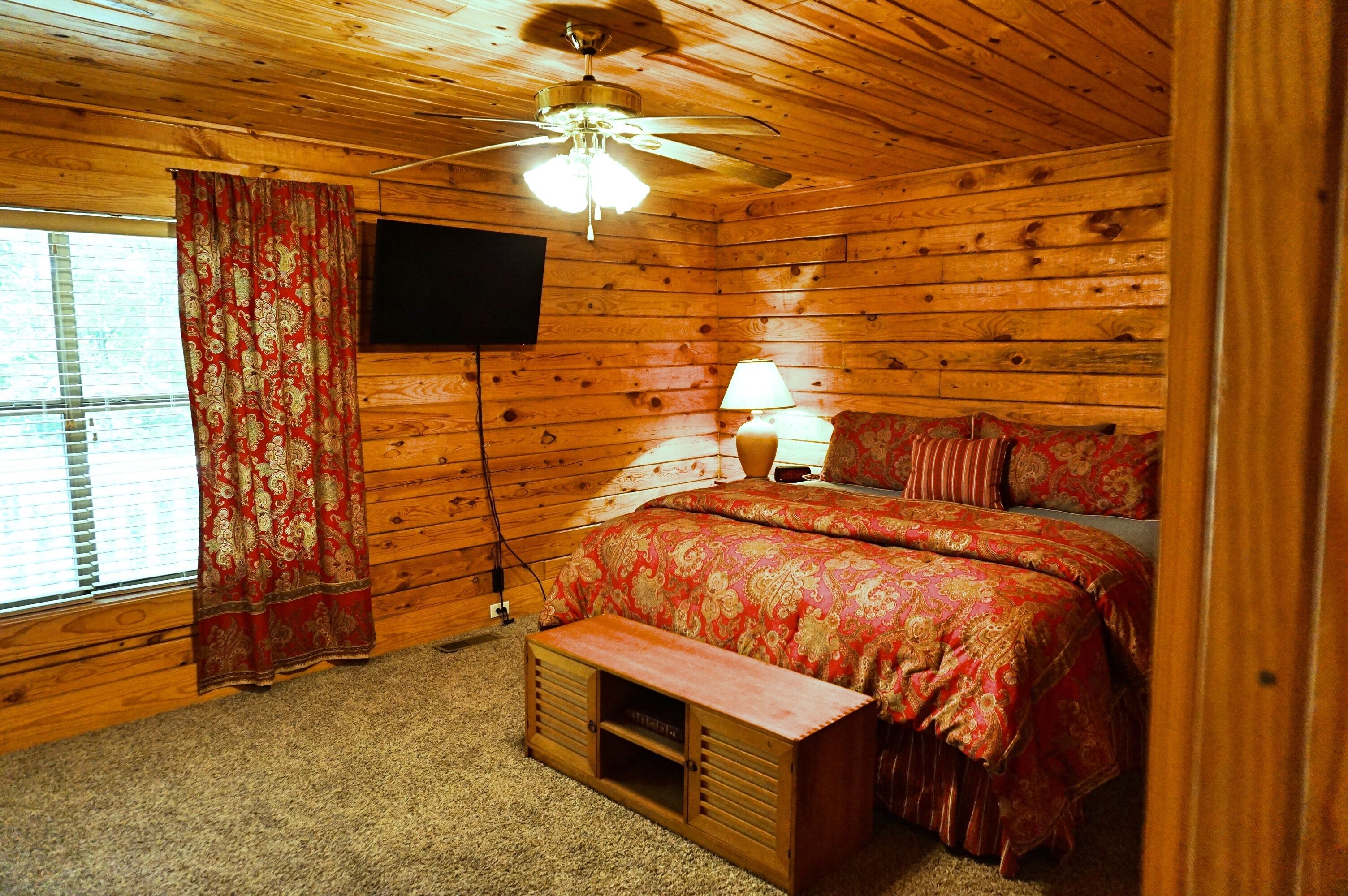 Une chambre à coucher aux murs lambrissés, avec un lit, un ventilateur de plafond, une télévision et une fenêtre avec des rideaux.