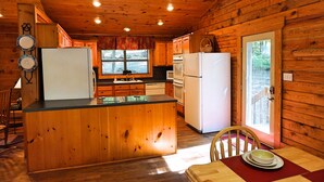 A wooden cabin interior with a kitchen featuring two refrigerators, a dining table with a red placemat, and a window overlooking a forested area.