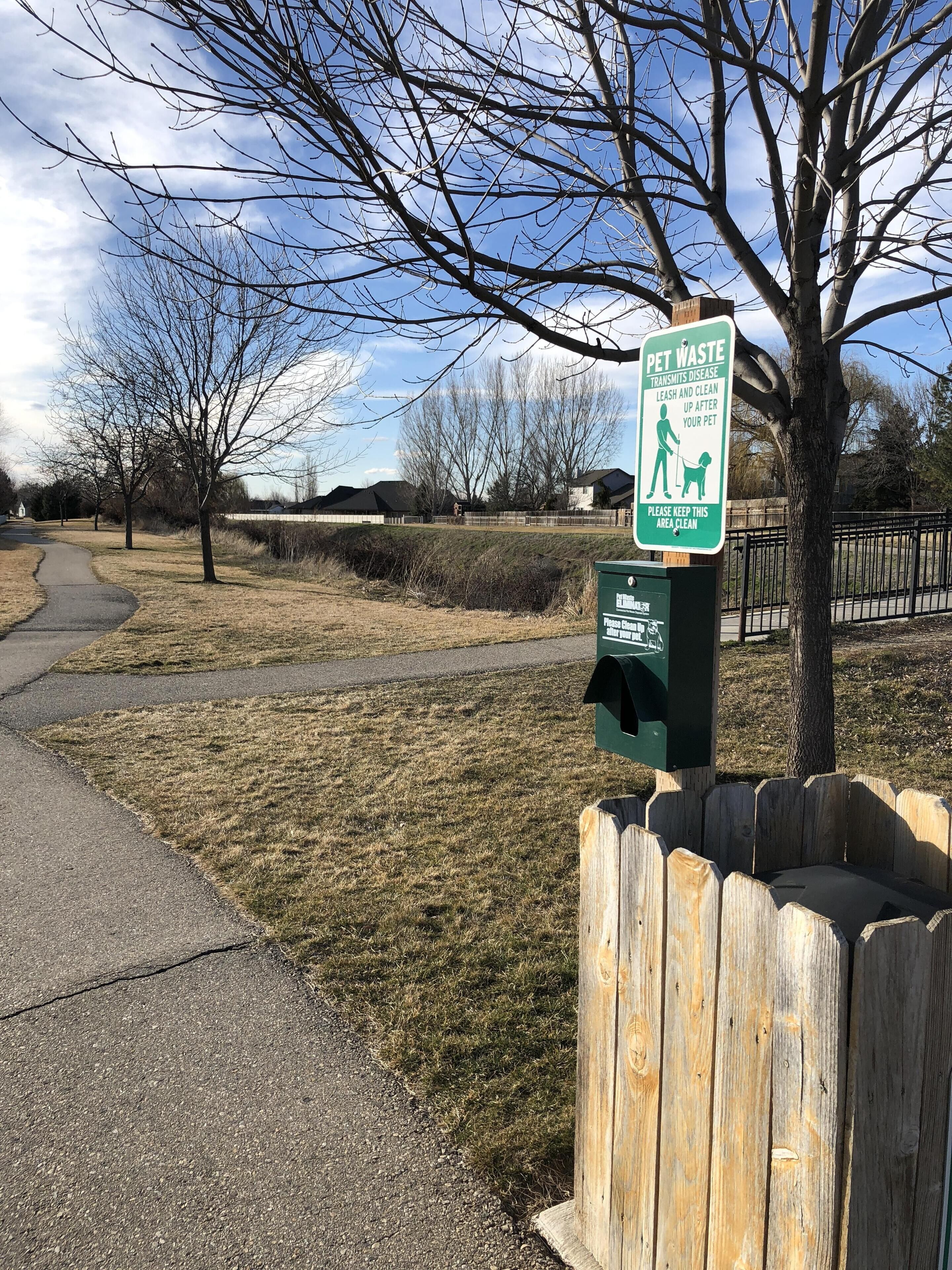 A park with a pet waste sign, a trash bin, and a wooden fence.