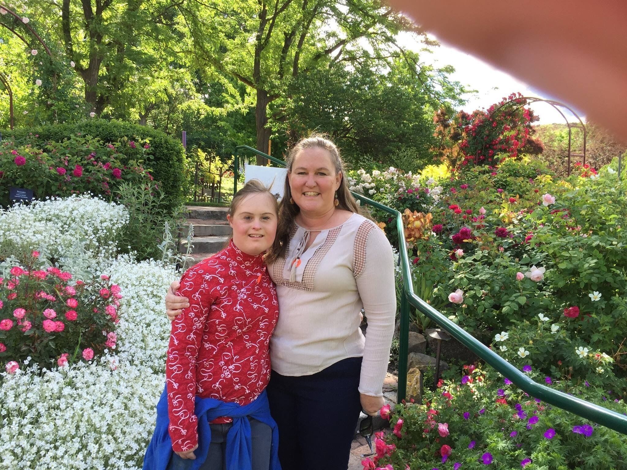 Une femme et un enfant se tiennent sur un escalier, entourés d’un jardin où poussent différentes sortes de fleurs.