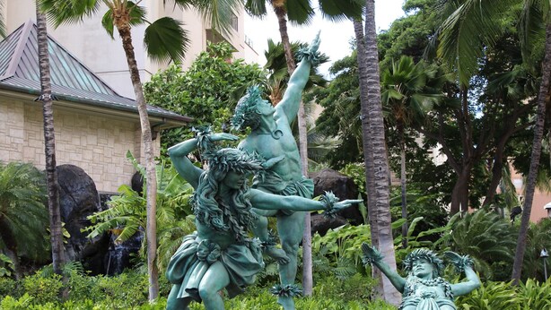 A bronze statue of a woman in a flowing dress and a man in a loincloth, set against a backdrop of lush greenery and a building with a tiled roof. - The Modern Suite -Waikiki Center Great Rates!! HK#112 (Honolulu)