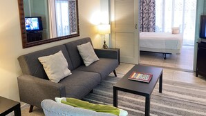 A living room with a sofa, a coffee table, a TV, and a bed in the adjacent room. - The Modern Suite -Waikiki Center Great Rates!! HK#112 (Honolulu)