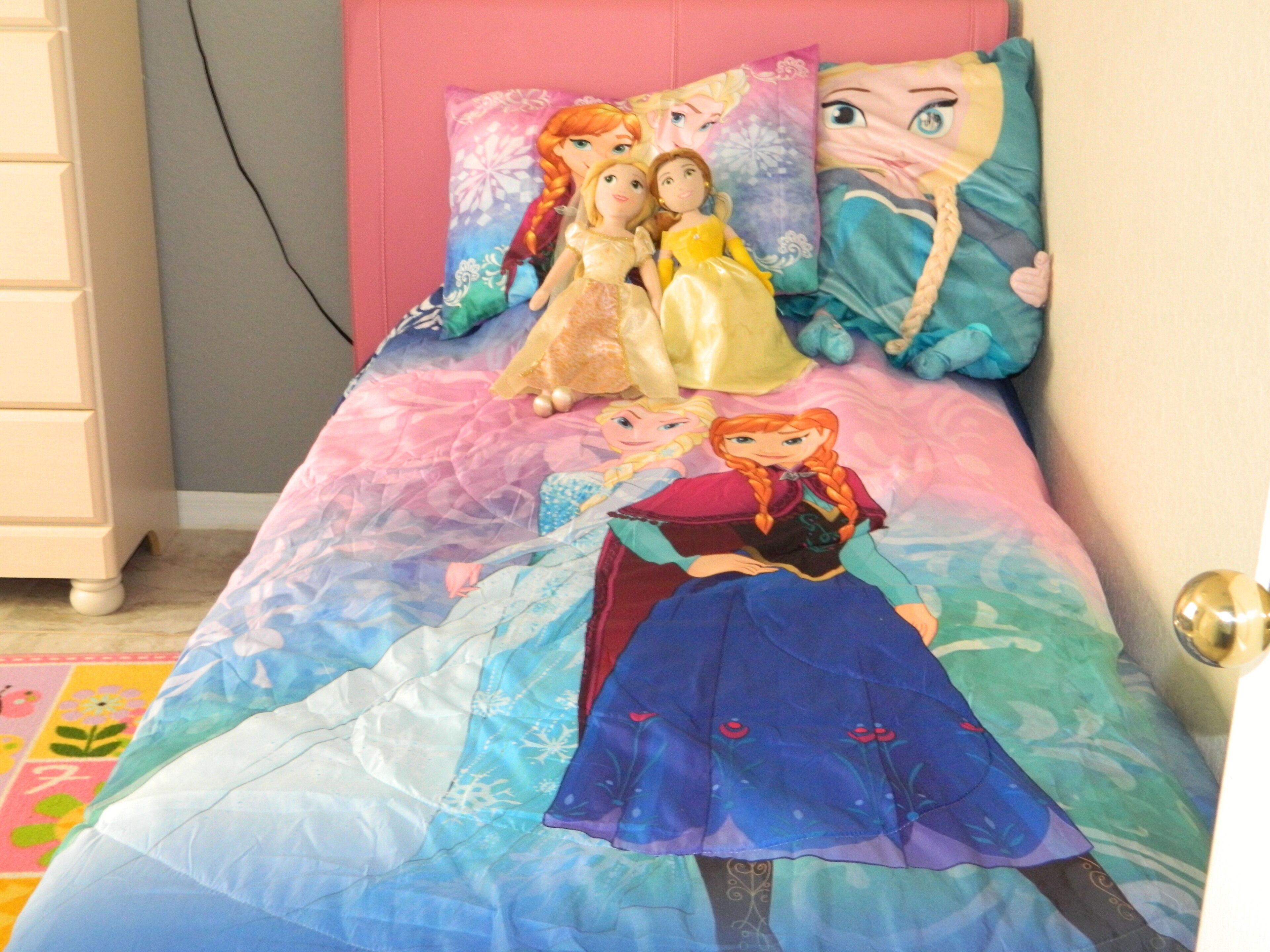 A bed with a colorful bedspread featuring Disney princesses, a pink headboard, and a white dresser with a mirror.