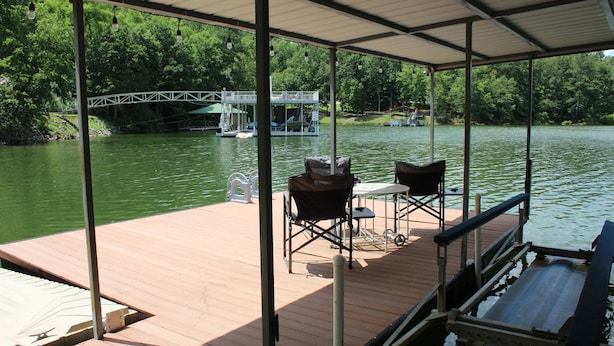 Een overdekte steiger met stoelen en een tafel, met uitzicht op een rustige rivier met een brug en steiger op de achtergrond.