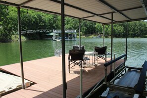 Un muelle techado con sillas y una mesa, con vista a un río tranquilo con un puente y un muelle al fondo.