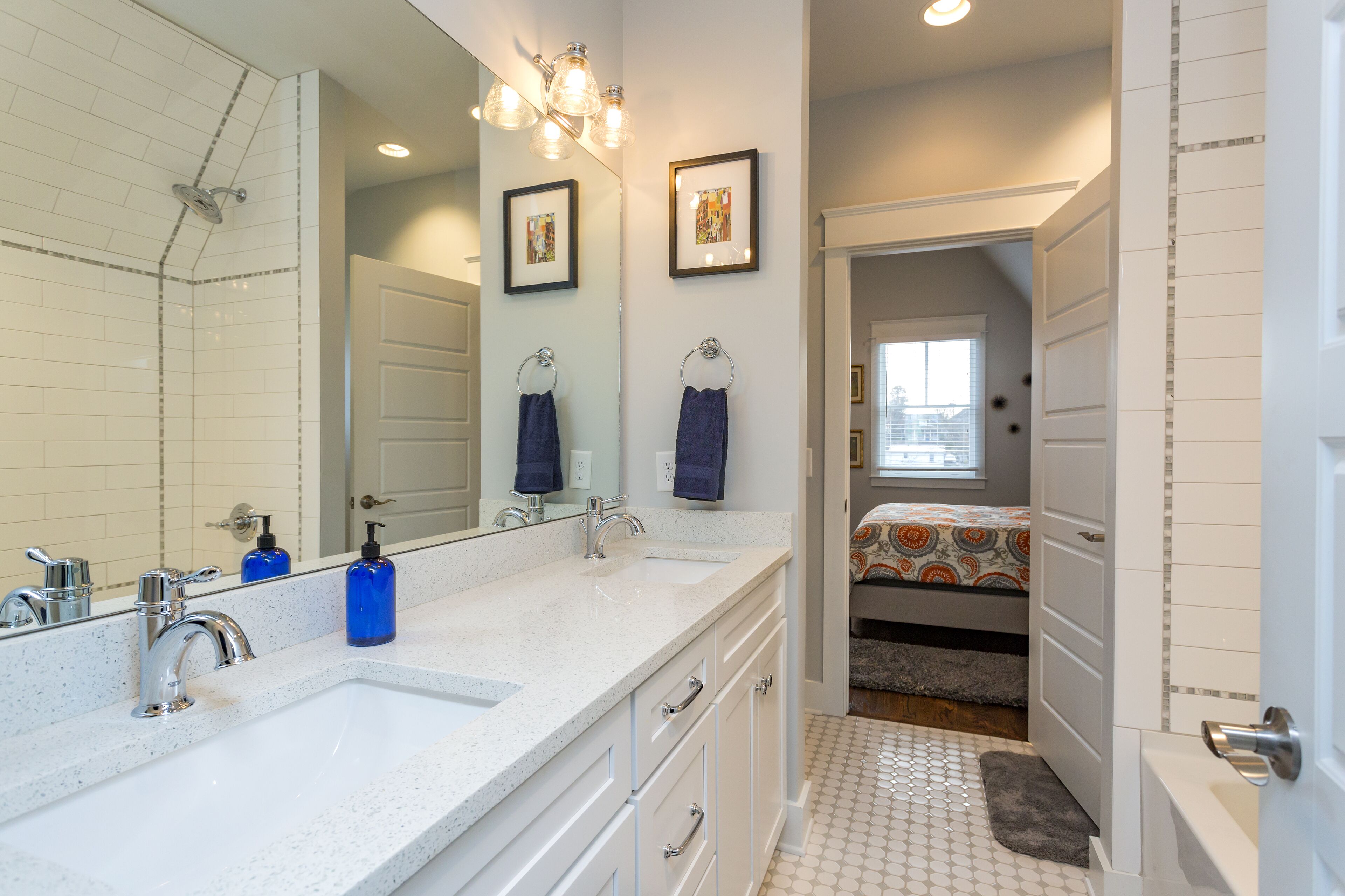 A bathroom with a double sink, a large mirror, and a shower. There is a bed visible through an open door in the background.