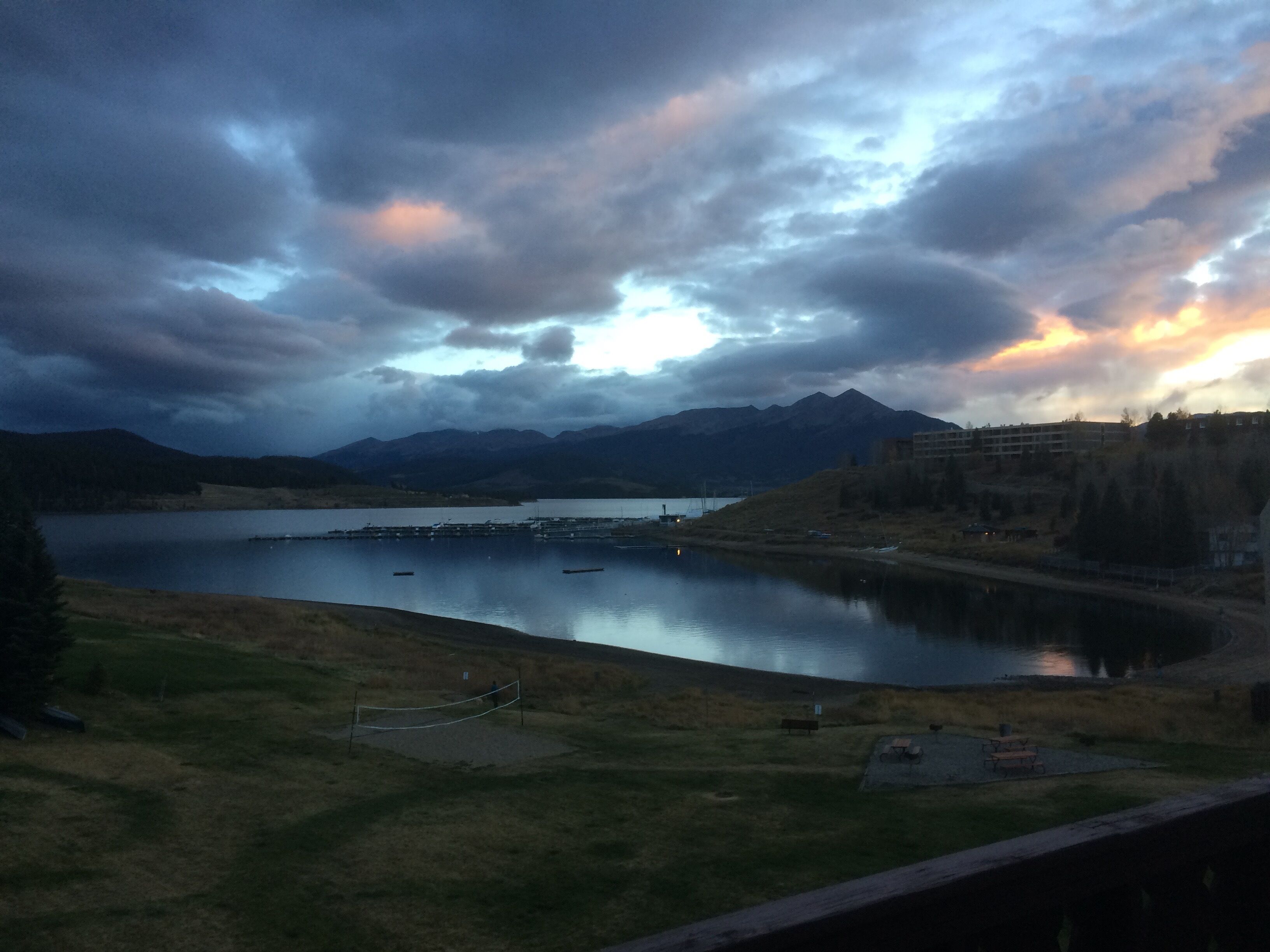 A lakeside view with mountains in the background, a dock, and a small building near the shore.