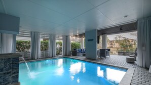 En indendørs swimmingpool med krystalklart udsyn til det omkringliggende område, inklusive et loungeområde med udendørs møbler og udsigt over bybilledet.