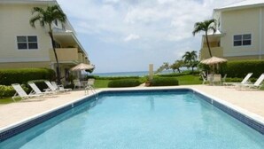 Piscina com espreguiçadeiras e guarda-sóis, cercada por edifícios com varandas e vista para o mar.