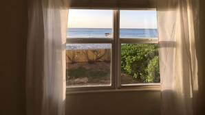 Uma janela com cortinas brancas, vista para a praia e alguma vegetação.