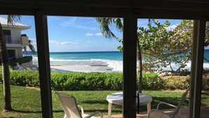 Pátio com mesa e cadeiras, vista para uma praia com ondas e céu claro.