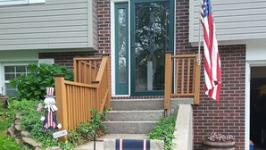 Ett hus med en fasad av tegel, en ytterdörr med ett glasparti, en veranda i trä, en amerikansk flagga och en välkomstskylt dekorerad med en basebollkeps.