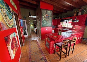 A kitchen with red tiled walls, a tiled floor, and a central island with chairs. There are paintings on the walls and a rug on the floor.