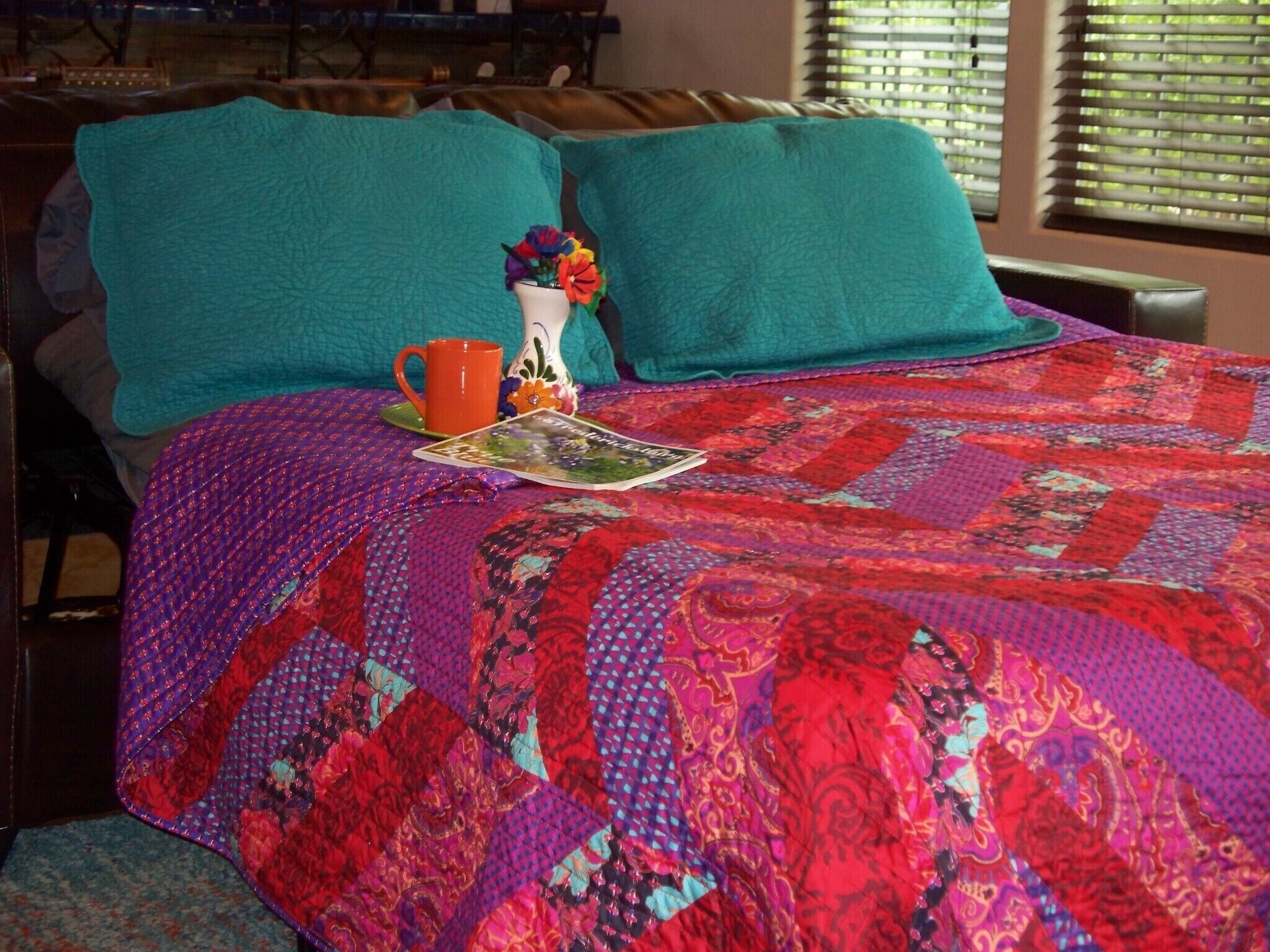 Una cama con un colcha colorida, almohadas color turquesa, un pequeño florero con flores y una taza roja.