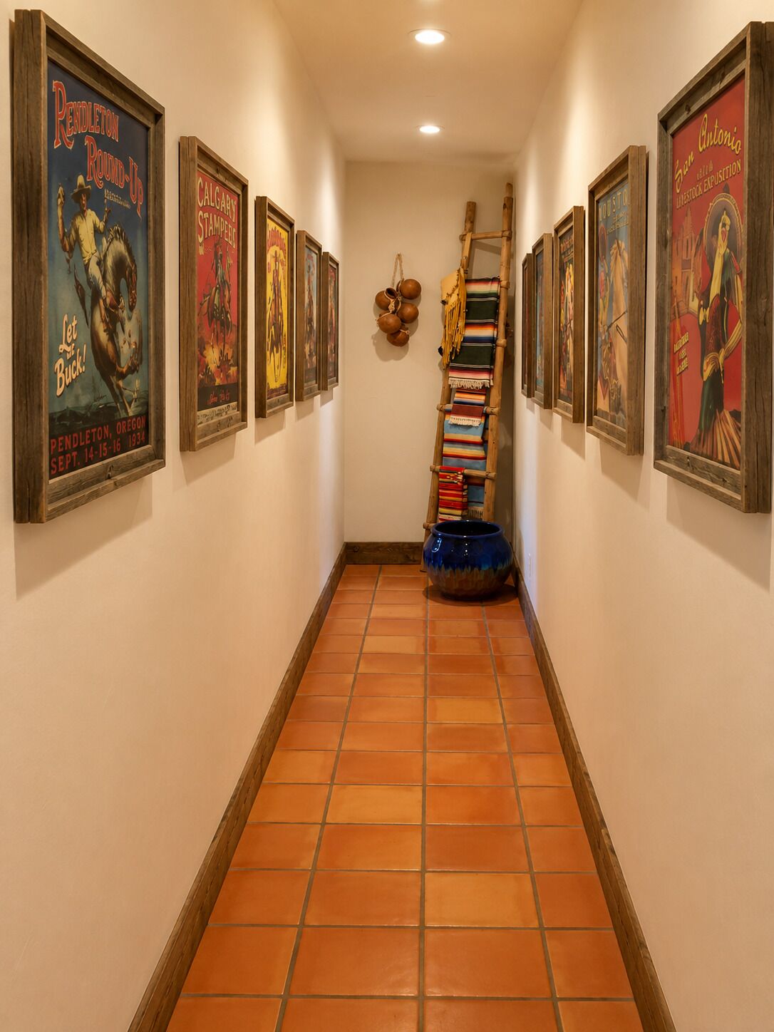 A hallway with framed posters on the walls, a wooden ladder leaning against the wall, and a blue pot on the floor.