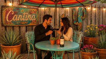 Deux personnes sont assises à une table ronde, une bouteille de vin devant elles, à l'ombre d'un parasol rayé aux couleurs vives. On aperçoit en arrière-plan une clôture en bois rustique et un panneau.