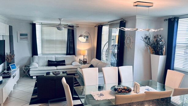 A modern living room with a glass dining table, white sofas, a TV, and a ceiling fan. - THE NAUTICAL!  FULLY EQUIPPED  3 BD 2BA APT AT AVENTURA  YACHT CLUB! (Aventura)