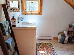 Un baño con tocador de madera, lavamanos, espejo, ventana, toallero, calefactor y alfombra estampada.