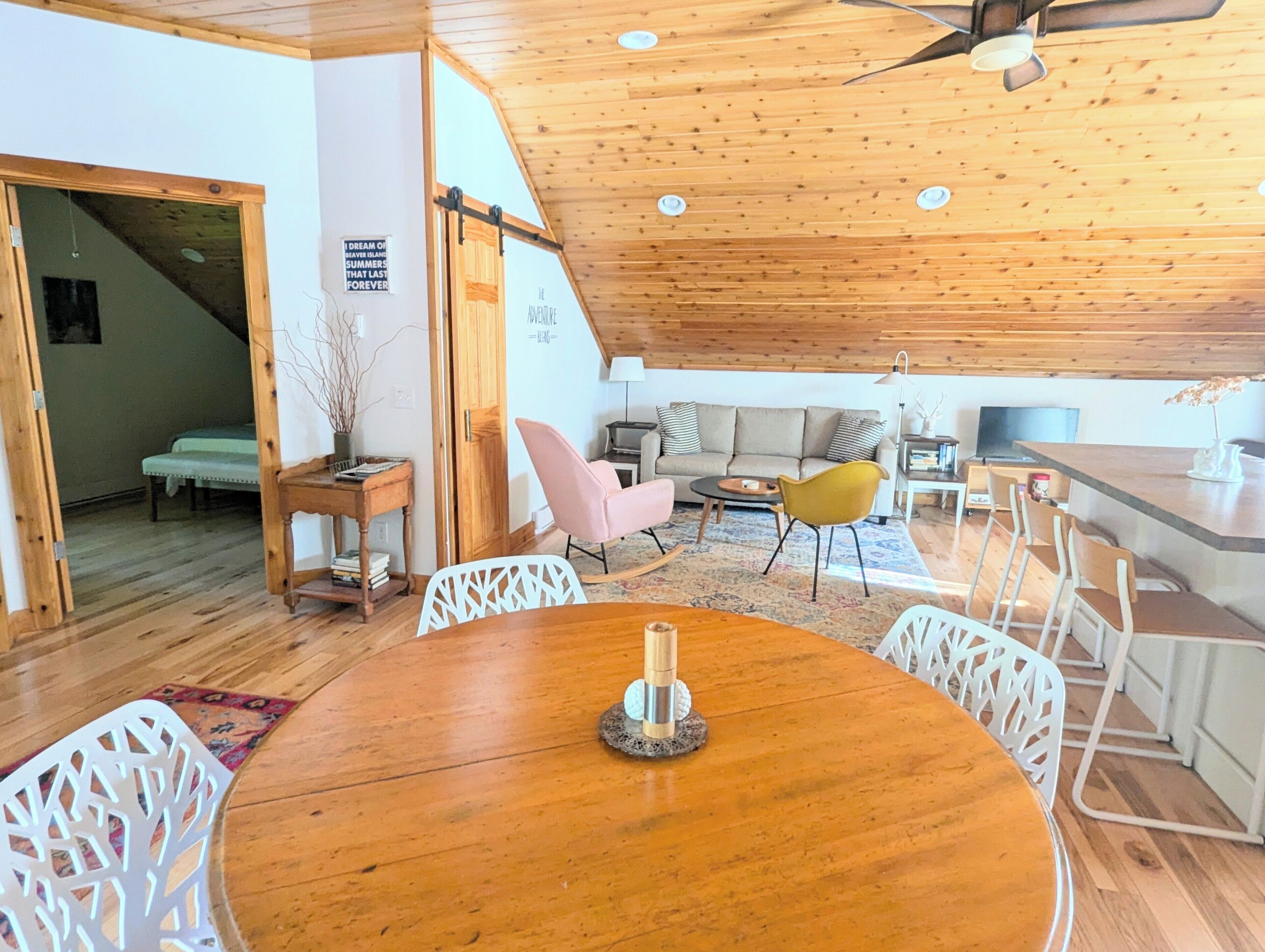 Un salon chaleureux avec un plafond en bois, une table à manger ronde entourée de chaises, un canapé et un téléviseur.